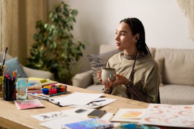 Genç bir Afrikalı Amerikalı kadın canlı sanat malzemeleriyle çevrili iken sıcak bir içkinin tadını çıkarıyor..