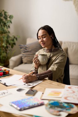 Saçları örgülü genç bir kadın, rahat dairesinde fikirler çizerken çayını yudumluyor, yaratıcılık saçıyor..