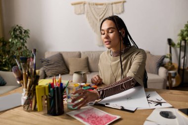 Güzel örgüleri olan genç siyah bir kadın sanatına odaklanmış, etrafı boyalarla çevrili..