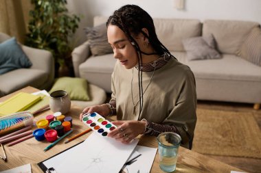 Saçları örülü genç bir kadın, şık dairesinde resim yapmaya dalmış, etrafı sanat malzemeleriyle çevrili..