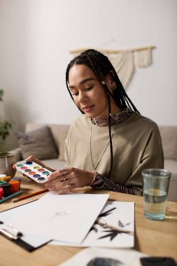 Saçları örgülü genç bir zenci kadın mutlu bir şekilde parlak ve modern dairesinde resim yapıyor..