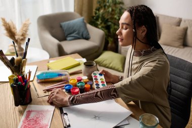 Saçları örgülü genç bir Afrikalı Amerikalı kadın şık evinde canlı renklerle resim yapmaktan hoşlanıyor..