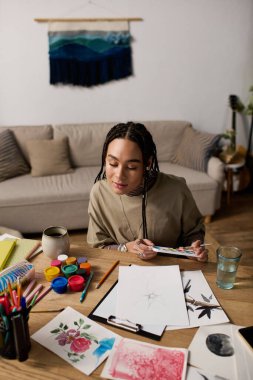 Genç bir Afrikalı Amerikalı kadın ahşap bir masada oturuyor, sanatına dalmış şık bir evde..