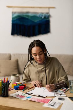 Örgülü genç siyahi bir kadın şık evinde bir masada sanat yaratıyor..