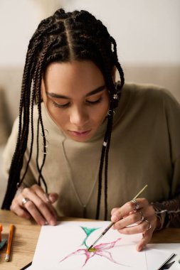 Saçları örgülü genç bir Afrikalı Amerikalı kadın rahat evinde canlı bir çiçek çizmeye odaklanıyor..