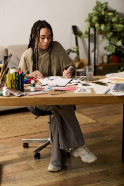 Saçları örgülü genç bir kadın, rahat dairesinde şık bir masada sanatsal çalışmalarına odaklanıyor..