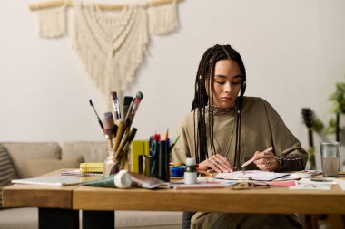 Evinde canlı sanat malzemeleriyle çevrili örgülü eskizler ve boyalarla genç bir kadın..