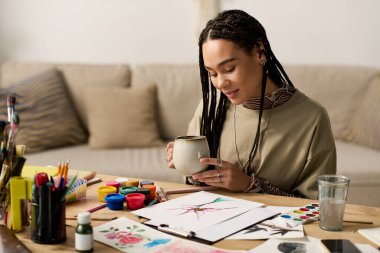 Şık giysiler içinde mutlu genç bir kadın davet ettiği yaşam alanında resim yaparken çayını yudumluyor..