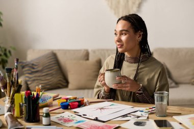 Genç bir kadın, etrafı sanat malzemeleriyle çevriliyken sıcak bir içecekle vakit geçirmekten zevk alır..