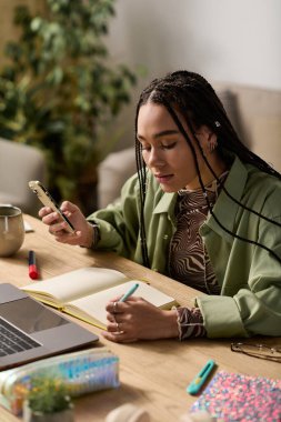 Saçları örgülü genç bir kadın rahat dairesinde fikirlerini yazarken telefonunu inceliyor..