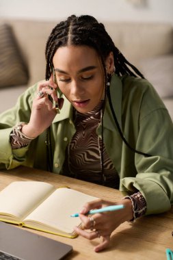 Saçları örgülü genç bir kadın telefon görüşmesi yapıyor. Moda evinde notlar yazıyor..