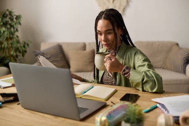 Süslü saçları olan genç siyah bir kadın şık bir mekanda dizüstü bilgisayarına odaklanırken kahvesini yudumluyor..