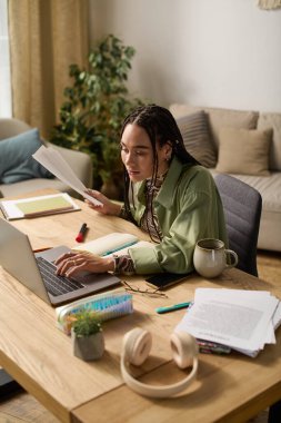 Örgülü genç bir Afrikalı Amerikalı kadın modern bir dairede masasında çalışıyor..