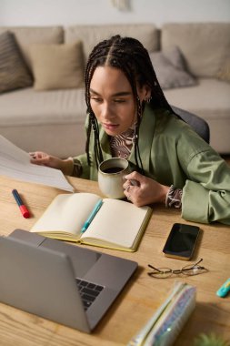 Modern bir dairede, örgülü genç siyah bir kadın çalışırken kahvesini yudumluyor..