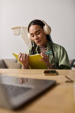 Saçları örgülü genç bir kadın, rahat dairesinde müzik dinlerken not alıyor..