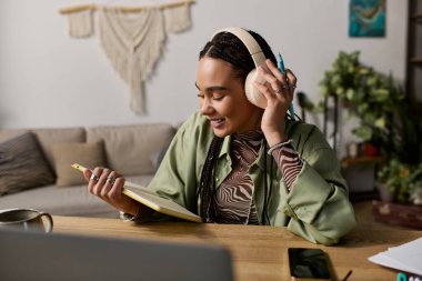 Saçları örgülü şık genç bir kadın güneşli dairesinde yazarken müzik dinliyor..