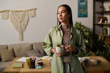Örgülü genç bir Afrikalı Amerikalı kadın modern dairesinde elinde fincanla duruyor..