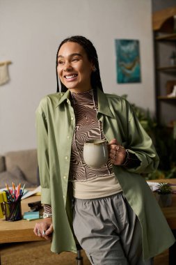 Saçları örgülü genç bir kadın evde kahvesinin tadını çıkarırken şık kıyafetler giyer..