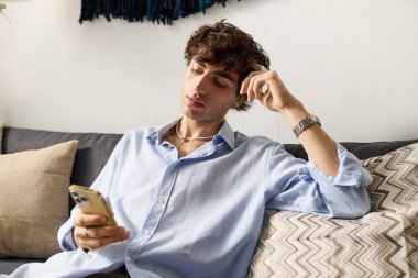 Kıvırcık saçlı genç bir adam modern dairesinde telefonunu kullanarak rahatlıyor..