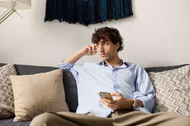 Kıvırcık saçlı şık bir genç adam şık dairesinde rahat bir şekilde oturuyor, telefonuyla meşgul..