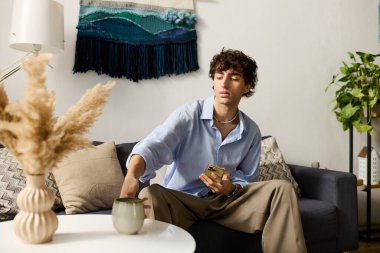 Kıvırcık saçlı genç bir adam modern dairesinde rahatça oturur, çayını yudumlar ve dinlenir..