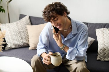 Yakışıklı, kıvırcık saçlı, elinde kahve kupasıyla telefonda gülümseyen genç bir adam..