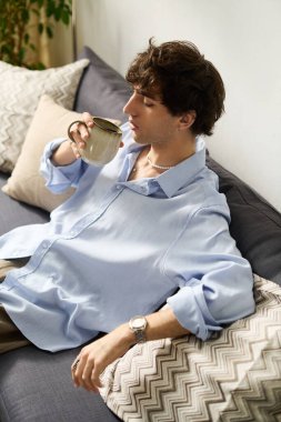 Mavi gömlekli genç adam bir kanepede dinleniyor, elinde bir fincan, evinde sakin bir anın tadını çıkarıyor..