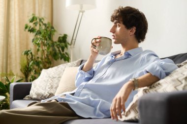 Kıvırcık saçlı yakışıklı genç bir adam kanepede rahatlar, rahat evinde bir fincan yudumlar..