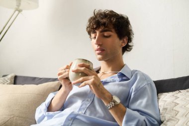 Kıvırcık saçlı genç adam zarif bir dairede sıcak bir içecekle rahatlıyor..