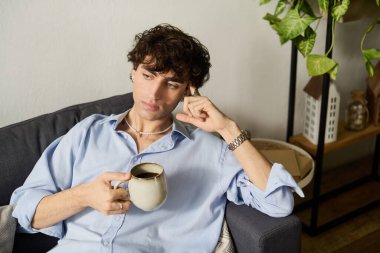 Kıvırcık saçlı yakışıklı genç bir adam modern bir dairede sessizce oturur ve kahvesini yudumlar..