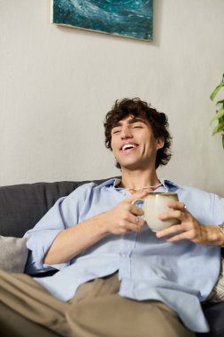 Kıvırcık saçlı genç bir adam rahat bir kanepede rahatlar ve son moda dairesinde kahvesini yudumlar..