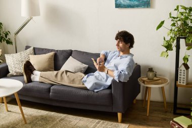 Kıvırcık saçlı genç bir adam rahat bir kanepede, telefonunda kayarak vakit geçirmekten hoşlanır..