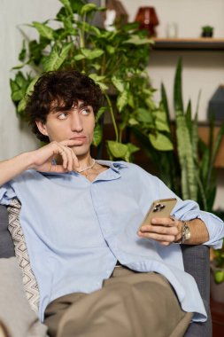Mavi gömlekli yakışıklı genç adam çiçeklerle çevrili modern bir dairede düşünceli bir şekilde oturuyor..