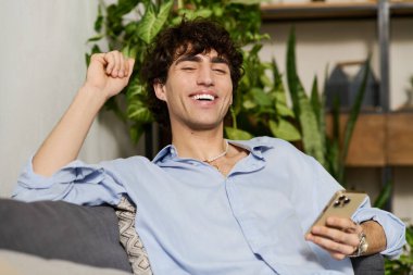Kıvırcık saçlı neşeli genç bir adam şık bir dairede koltukta dinleniyor ve telefonunun keyfini çıkarıyor..