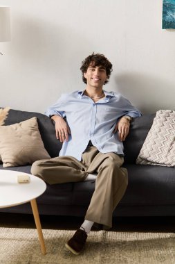 Yakışıklı genç bir adam, modern dairesinde, mavi gömlek ve pantolon giyerek aylak aylak dolaşıyor..