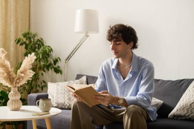 Çekici bir genç adam modern dairesinde rahat bir şekilde oturuyor, bir kitaba dalmış..