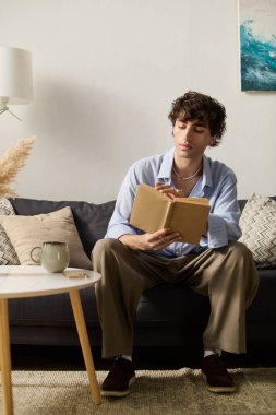 Kıvırcık saçlı genç bir adam şık dairesinde kitap okuyor.