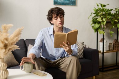Kıvırcık saçlı yakışıklı bir genç adam modern dairesinde rahatça oturur ve kitap okur..