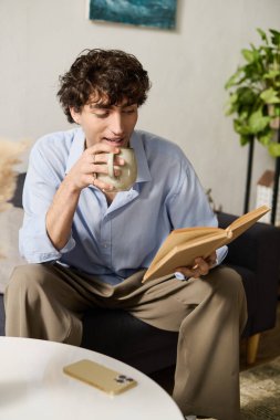 Kıvırcık saçlı genç adam, rahat, modern evinde kitap ve kahveyle rahatlıyor..