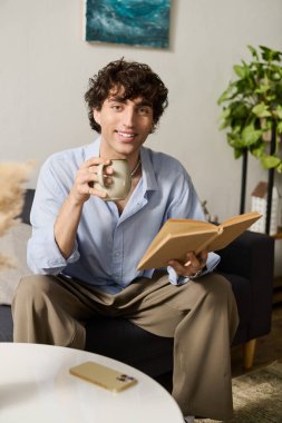 Neşeli genç bir adam modern dairesinde oturuyor, kitap okurken fincandan yudumluyor..