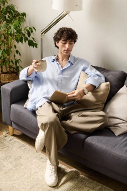 Modern dairesinde rahat bir genç adam kitap okurken çayını yudumluyor..