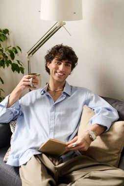 Kıvırcık saçlı genç bir adam modern bir dairede elinde bir bardak ve defterle rahatça oturuyor..