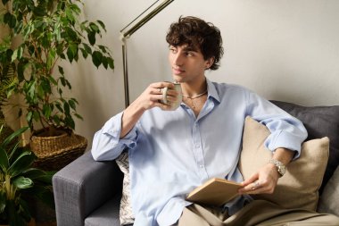 Yakışıklı genç bir adam bir kanepede dinleniyor, bitkilerle çevrili olarak kahvenin tadını çıkarıyor..