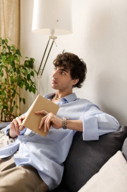 Kıvırcık saçlı genç bir adam modern bir dairede oturuyor ve kitabı yansıtıyor..