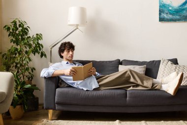 Genç bir adam rahat bir şekilde kanepede uzanıyor, modern dairesinde kitap okumaya dalmış..