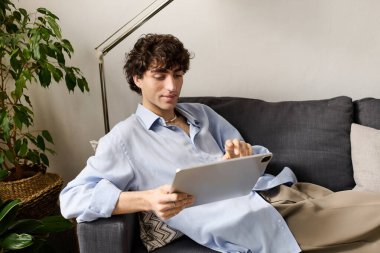 Kıvırcık saçlı genç adam tabletiyle uğraşırken şık dairesinde rahatlıyor..