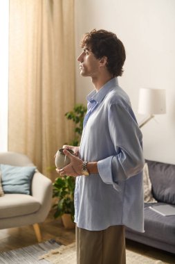 Yakışıklı bir genç adam zarif dairesinde elinde bir kupayla düşünceli bir şekilde duruyor..