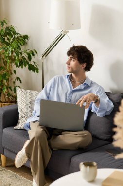 Kıvırcık saçlı genç adam koltuğunda dinleniyor ve şık dairesinde dizüstü bilgisayarla vakit geçiriyor..