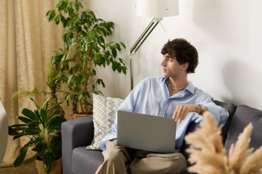 Kıvırcık saçlı genç bir adam, modern dairesinde dizüstü bilgisayarıyla rahat bir koltukta oturur, düşüncelere dalmıştır..