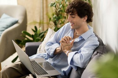 Kıvırcık saçlı genç bir adam modern dairesinde bir kanepede dizüstü bilgisayarla oturuyor..